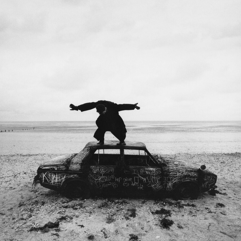 Cover of an album by The 1975, Being Funny in a Foreign Language. Black and white photograph of a person standing on a burnt out car at the beach.